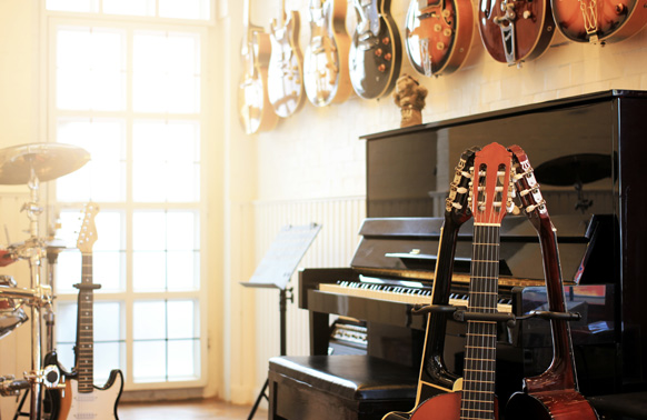 A music room with several guitars hanging on the wall. A drum kit and a piano are in the midground. Three guitars in stands are arranged together close to the piano in the forground. 