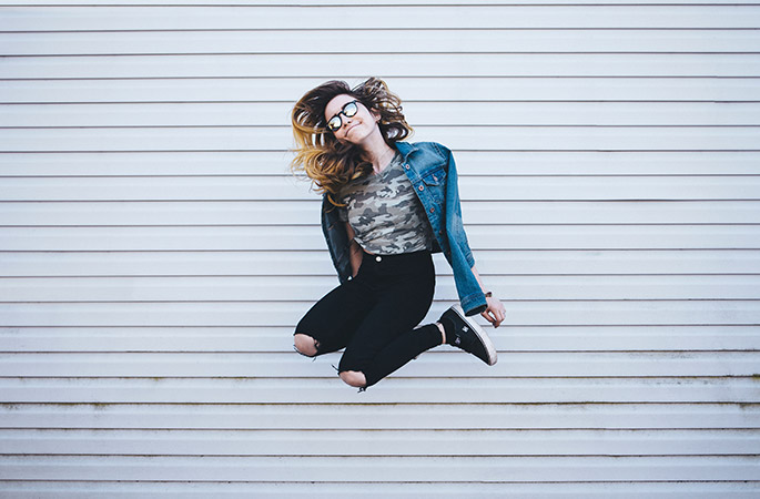 Woman with denim jacket and reflective glasses jumping in the air.