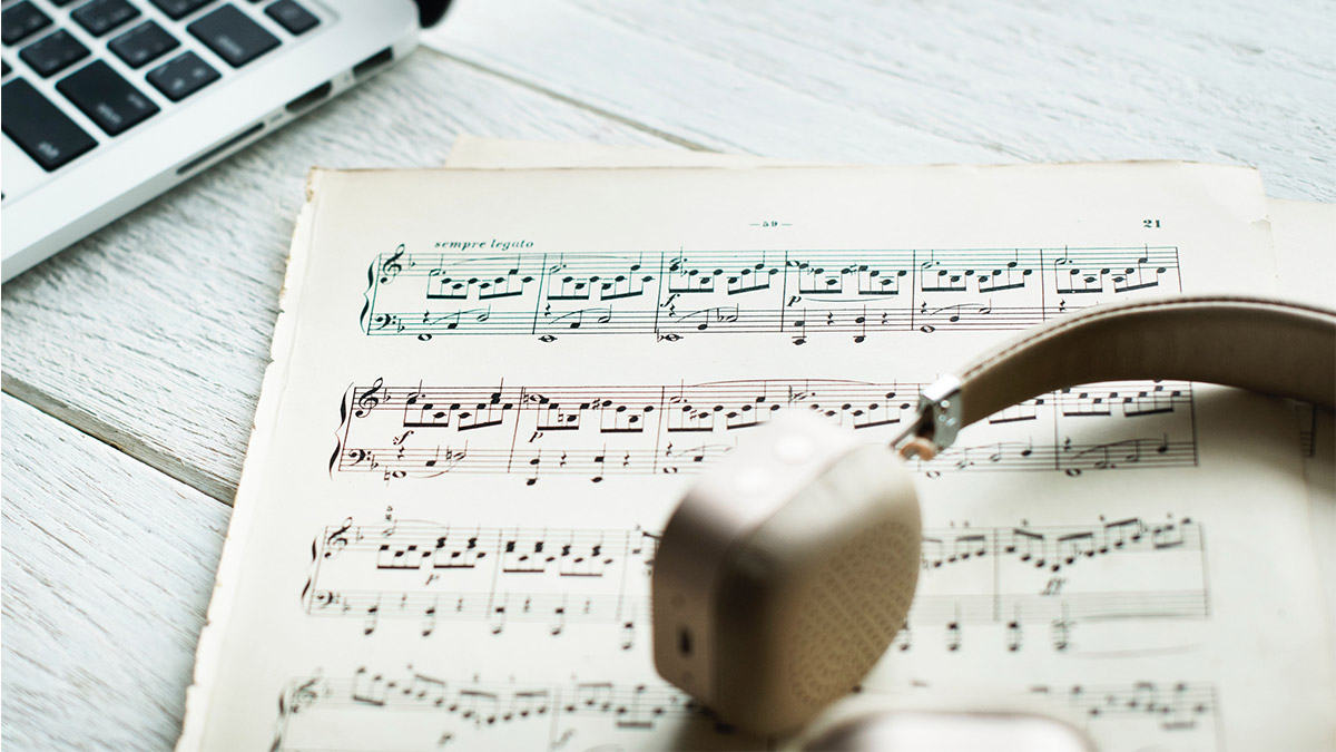 Music book open on a wooden table. Over-the-ear headphones are laying on top of the book, and a laptop is in the background.